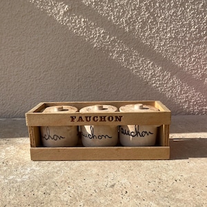 Pode incluir: Um conjunto de três pequenos potes de cerâmica em tom esbranquiçado com tampas, cada um apresentando o texto cursivo preto Fauchon, alojados numa caixa de madeira retangular com o nome da marca Fauchon marcado em letras escuras na ripa frontal.