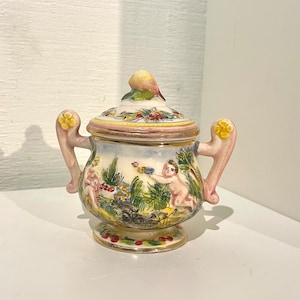 Può includere: Una piccola zuccheriera in ceramica bianca con coperchio. La ciotola è decorata con una scena colorata di cherubini che giocano in un giardino. Il coperchio ha un piccolo uccello come pomo.