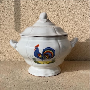 Può includere: Zuppiera in ceramica bianca con coperchio, decorata con un gallo dipinto a mano con accenti blu, rossi, gialli e verdi. La zuppiera ha due manici e una base arrotondata.