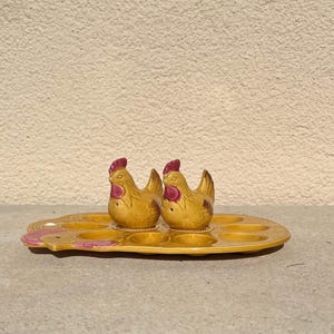 May include: A ceramic egg holder shaped like a chicken with six individual egg cups. Two small ceramic chickens with red combs are perched on the holder.