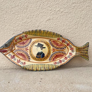 Könnte beinhalten: Eine Keramik-Servierplatte in Fischform mit einem farbenfrohen Design. Die Platte zeigt ein zentrales Bild einer Person mit Hut und einem blau-weiß gemusterten Hemd. Die Platte ist mit roten, gelben und blauen Blumenmustern verziert.