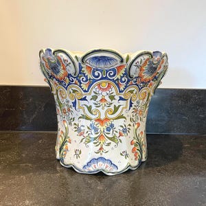 Pode incluir: Um vaso de cerâmica branco com uma borda ondulada e um design floral em azul, amarelo, laranja e verde. O vaso é decorado com uma variedade de flores e folhas.