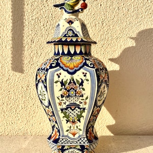 Könnte beinhalten: Eine große, blaue und weiße Keramikvase mit einem Vogelaufsatz. Die Vase ist mit floralen Mustern und geometrischen Designs in Blautönen, Gelb, Orange und Grün verziert.