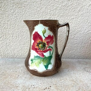 May include: A brown ceramic pitcher with a floral design. The pitcher features a red poppy flower with green leaves and a brown background.