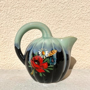 May include: A small, green and black ceramic pitcher with a floral design. The pitcher has a round body and a curved handle. The floral design features red poppies, white daisies, and blue flowers.