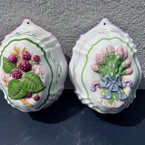 May include: Two white ceramic wall plaques with scalloped edges. One features a raspberry and floral design, the other a bundle of asparagus tied with a ribbon. Green and pink accents.