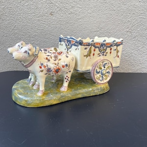 Könnte beinhalten: Eine weiße Keramikfigur von zwei Kühen, die einen kleinen Wagen ziehen. Der Wagen und die Kühe sind mit bunten Blumenmustern und blauen und grünen Akzenten verziert.