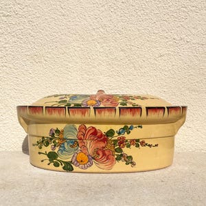 Può includere: Una casseruola ovale in ceramica gialla con un motivo floreale. I fiori sono dipinti in tonalità di rosa, blu, giallo e verde. La casseruola ha un bordo rosso e un piccolo pomo sul coperchio.
