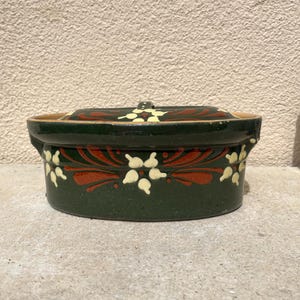 Pode incluir: Uma travessa de cerâmica verde escura com tampa. O prato é decorado com padrões florais vermelhos e creme. A travessa está sobre uma superfície de concreto.