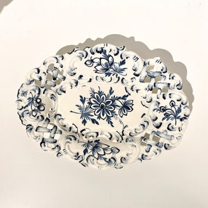 Peut inclure: Un plat de service ovale en céramique blanche avec un motif floral bleu et blanc. Le plat a un design délicat et ajouré autour du bord.