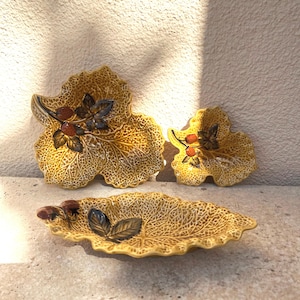 May include: Set of three decorative leaf-shaped serving dishes. The dishes are a textured yellow with brown and black accents, featuring a leaf and berry design. The set includes two larger leaf-shaped dishes and one smaller, elongated dish.