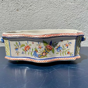 Könnte beinhalten: Ein weißer Keramik-Pflanztopf mit einem gewellten Rand und blauen, roten und grünen Blumendesigns. Der Topf hat zwei kleine Füße auf jeder Seite.