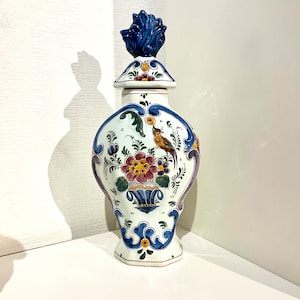 Könnte beinhalten: Eine weiße Keramikvase mit einem blauen und weißen Blumendesign und einem Vogel. Die Vase hat einen Deckel mit einem blauen und weißen Blumendesign.