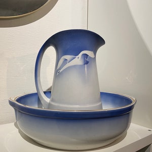 May include: A blue and white ceramic pitcher with a swan design, set inside a matching bowl. The pitcher has a curved handle and a wide spout. The bowl is round with a slightly flared rim. The set is displayed on a white shelf.