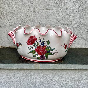 Op de afbeelding: Een witte keramische kom met een gegolfde rand en een rood bloemenontwerp. De kom heeft twee handvatten en is versierd met rode, roze en groene bloemen.
