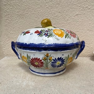 May include: A white ceramic serving dish with a lid, featuring a blue and white floral design with yellow and red accents. The dish has two handles and a round base.
