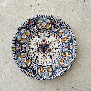 Könnte beinhalten: Ein weißer Keramikteller mit einem blauen, grünen, orangefarbenen und roten Blumendesign. Der Teller hat einen gewellten Rand und Löcher am Rand.