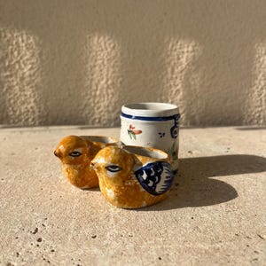 May include: Two small ceramic bird-shaped egg holders with blue and white painted designs. A small white ceramic mug with blue and orange painted designs sits behind the egg holders.