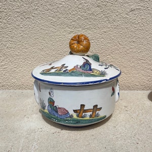 May include: A white ceramic covered dish with blue and green painted details. The lid features a small orange pumpkin-shaped knob. The dish is decorated with a scene of two people sitting on a bench.