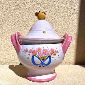 Könnte beinhalten: Eine weiße Keramik-Servierplatte mit einem rosafarbenen Deckel und rosafarbenen Griffen. Die Platte ist mit rosa Blumen und blauen Schmetterlingen verziert.