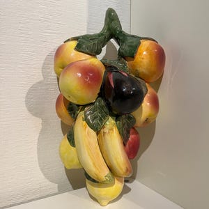 Puede incluir: Una decoración de pared de cerámica con un racimo de frutas, incluyendo plátanos amarillos, manzanas rojas, una berenjena morada y limones amarillos. La fruta está dispuesta en una vid verde con hojas.