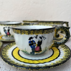 Könnte beinhalten: Ein Set vintage Teetassen und Untertassen mit gelbem und schwarzem Rand. Die Tassen zeigen ein handgemaltes Design eines Paares in traditioneller Kleidung.