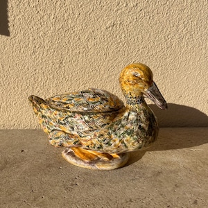 May include: A ceramic duck figurine with a lid. The duck has a yellow head and beak, with a body featuring a mix of green, yellow, and brown patterns. The base is a light yellow color. The figurine is sitting on a gray surface.