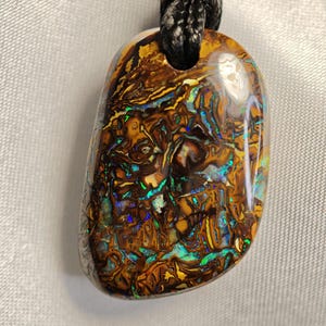 Op de afbeelding: Een ketting met een hanger van een grote, onregelmatig gevormde opaal. De opaal is een levendige mix van blauwe, groene, gouden en bruine kleuren. De steen is gezet in een zilveren of gouden setting en hangt aan een zwart koord.