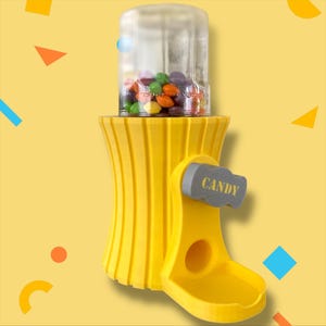 Peut inclure: Un distributeur de bonbons jaune avec un dessus transparent rempli de bonbons colorés. Le distributeur a une poignée grise avec le mot "CANDY" imprimé dessus. Le distributeur est sur un fond jaune.