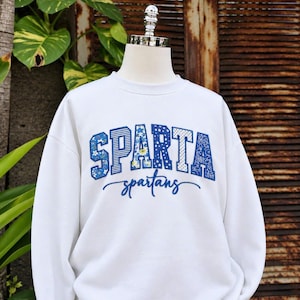 May include: A white crewneck sweatshirt featuring the word SPARTA in large, blue-toned patterned block letters, with the word spartans written in a blue cursive script underneath. The patterns on the block letters include stripes, florals, and stars.