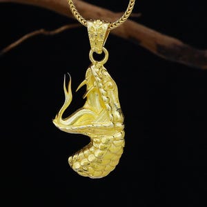 Könnte beinhalten: Eine goldfarbene Halskette mit einem detaillierten Schlangenanhänger. Der Kopf der Schlange ist geöffnet und zeigt ihre Zähne. Der Anhänger hängt an einer filigranen Goldkette vor dunklem Hintergrund.