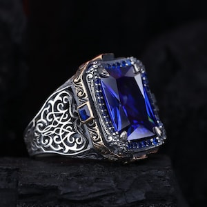 Puede incluir: Anillo de plata con una gran gema rectangular de color azul zafiro. El anillo presenta intrincados detalles de filigrana en los laterales y un borde dorado. El anillo se muestra sobre un fondo oscuro.