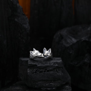 May include: A silver ring featuring two detailed fox heads facing each other. The ring is set against a dark, textured background, highlighting the intricate design and metallic sheen of the jewelry. The fox heads are sculpted with realistic features.
