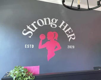 May include: A wall decal featuring the words "Strong HER" in white, with a pink silhouette of a woman lifting a weight. The text "ESTD 2020" is also present. The decal is on a gray wall.