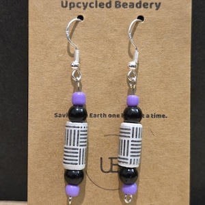 May include: A pair of handmade earrings with silver hooks. Each earring features a purple bead, a black bead, a white and black patterned bead, another black bead, and a final purple bead. The earrings are displayed on a brown card with the text "Upcycled Beadery".
