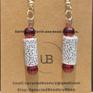 May include: A pair of handmade earrings featuring a silver chain, red beads, and white cylindrical beads with a black pattern. The earrings have a unique design and are displayed against a brown background. The text "Saving Earth one bead" is visible.