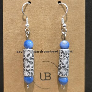 May include: A pair of dangle earrings with silver hooks. Each earring features a blue bead, a white and black patterned cylinder bead, and another blue bead. The earrings are displayed on a brown card with text.