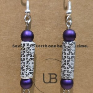 May include: A pair of dangle earrings with silver-tone hooks. Each earring features a purple bead, a white cylinder with a black floral pattern, and another purple bead. The earrings are displayed against a brown background.