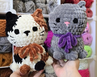 May include: Two handmade crochet cat plushies. One is gray with a purple bow, the other is calico with black, brown, and white. Both have black eyes and pink noses. The plushies are displayed in front of a shelf filled with yarn.