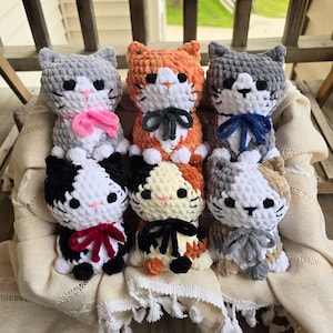 May include: Six plush crochet cat toys arranged in two rows. The cats feature soft yarn textures, black bead eyes, pink stitched noses, and small bows in various colors including pink, dark gray, blue, red, and black, all sitting in a cream fabric.