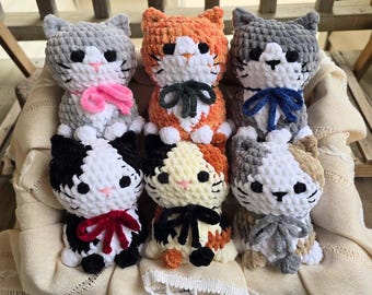 May include: Six plush crochet cat toys arranged in two rows. The cats feature soft yarn textures, black bead eyes, pink stitched noses, and small bows in various colors including pink, dark gray, blue, red, and black, all sitting in a cream fabric.