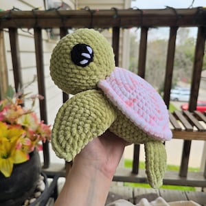 May include: A plush turtle toy crafted from soft, textured yarn. The head and flippers are a solid olive green, while the shell features a variegated pattern of white and soft pink. The turtle has large, round black eyes with white highlights.