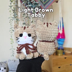 May include: Two light brown tabby cat plush toys made of soft yarn. The front-facing cat has a white chest, black eyes, a pink nose, and a dark brown ribbon tied around its neck. The text Light Brown Tabby is displayed above the plush toys.