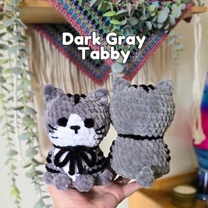 May include: Two plush cat toys made of dark gray and white yarn. The front-facing cat has a white face, black eyes, a black nose, and a black bow. The back-facing cat is solid dark gray with black stripes. Text above reads: Dark Gray Tabby.