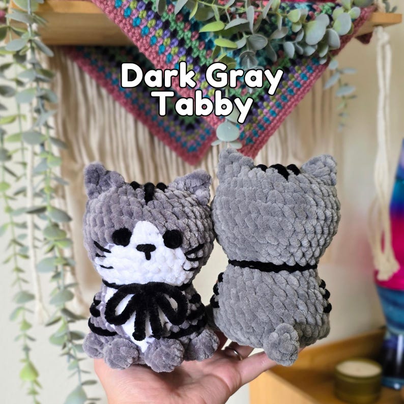 May include: Two plush cat toys made of dark gray and white yarn. The front-facing cat has a white face, black eyes, a black nose, and a black bow. The back-facing cat is solid dark gray with black stripes. Text above reads: Dark Gray Tabby.