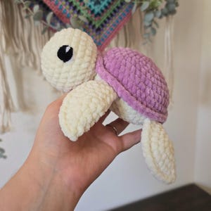 May include: A handmade crochet turtle plushie in cream, lavender, and black. The turtle has a cream-colored head and limbs, a lavender shell, and a black eye. The plushie is soft and textured, perfect for a child's toy or nursery decor.