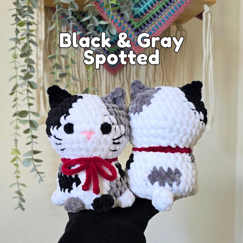 May include: Two handmade crochet cat plushies featuring black, gray, and white yarn. The front-facing cat has a pink nose, black eyes, and a red ribbon tied in a bow. The second cat is shown from the back with a red collar. Text reads Black & Gray Spotted.