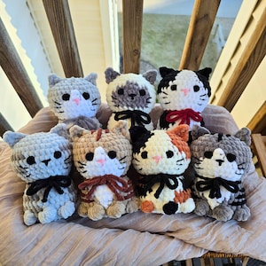 May include: Seven small, handmade crochet cat plushies arranged in two rows. Each cat features a soft, chunky yarn texture, black button eyes, a small pink stitched nose, and whiskers. Several cats wear small fabric bows around their necks.