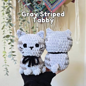 May include: Two gray striped tabby cat plush toys made of soft yarn. The cat on the left features black embroidered eyes, a nose, whiskers, and a black ribbon tied around its neck. The text Gray Striped Tabby is visible above the plush toys.