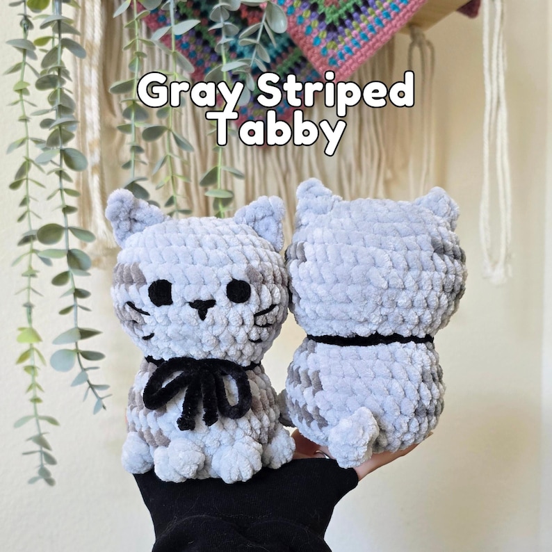 May include: Two gray striped tabby cat plush toys made of soft yarn. The cat on the left features black embroidered eyes, a nose, whiskers, and a black ribbon tied around its neck. The text Gray Striped Tabby is visible above the plush toys.